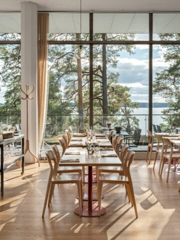 Bilden förställer Artipelag Restaurang i solljus. På bilden syns dukade bord på rad framför ett panoramafönster med utsikt över vattnet och talltopparna.