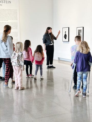 Bild på en konstvärd som håller i en konstvisning för barn. I bilden syns en grupp barn som står vända mot konstvärden. I bakgrunden skymtar två konstverk.